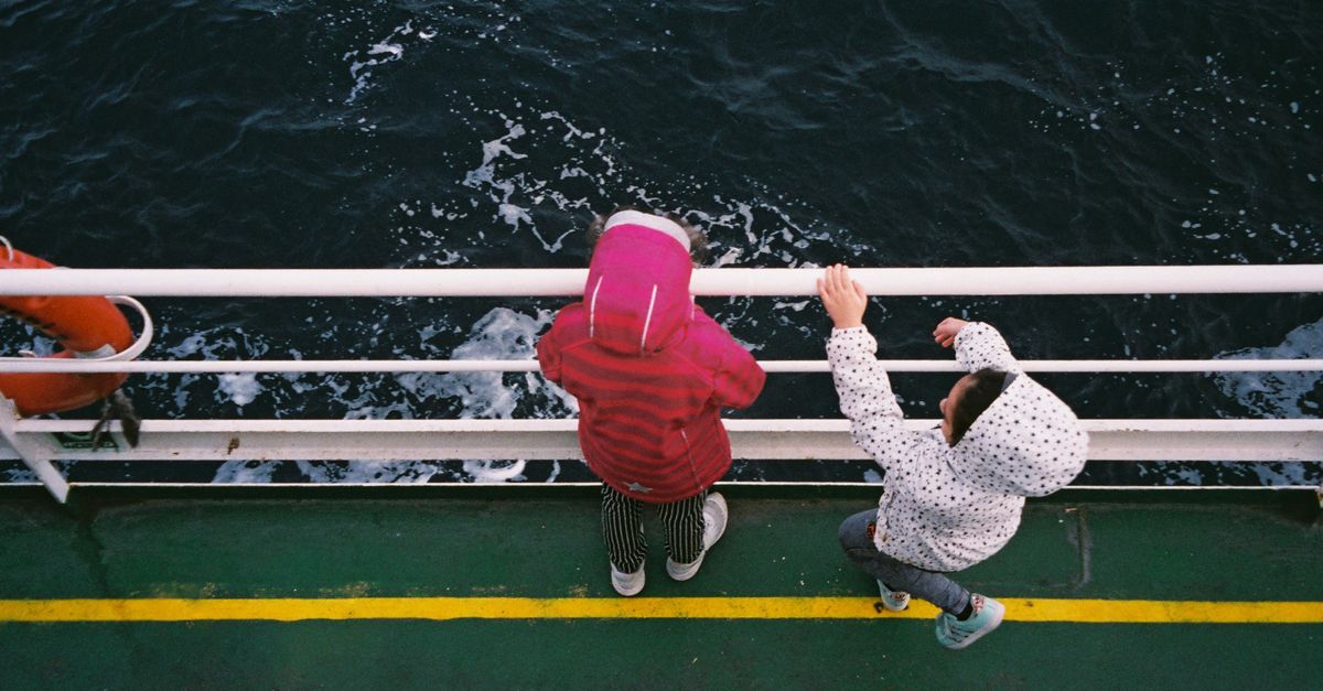 Two children in jackets standing at a boat railing, looking out across the sea