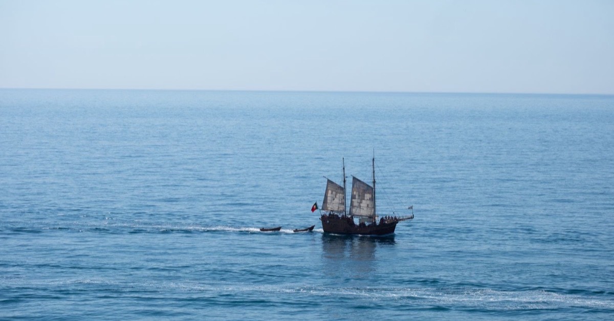 A sailing vessel with dark sails towing two small tenders across the open Atlantic