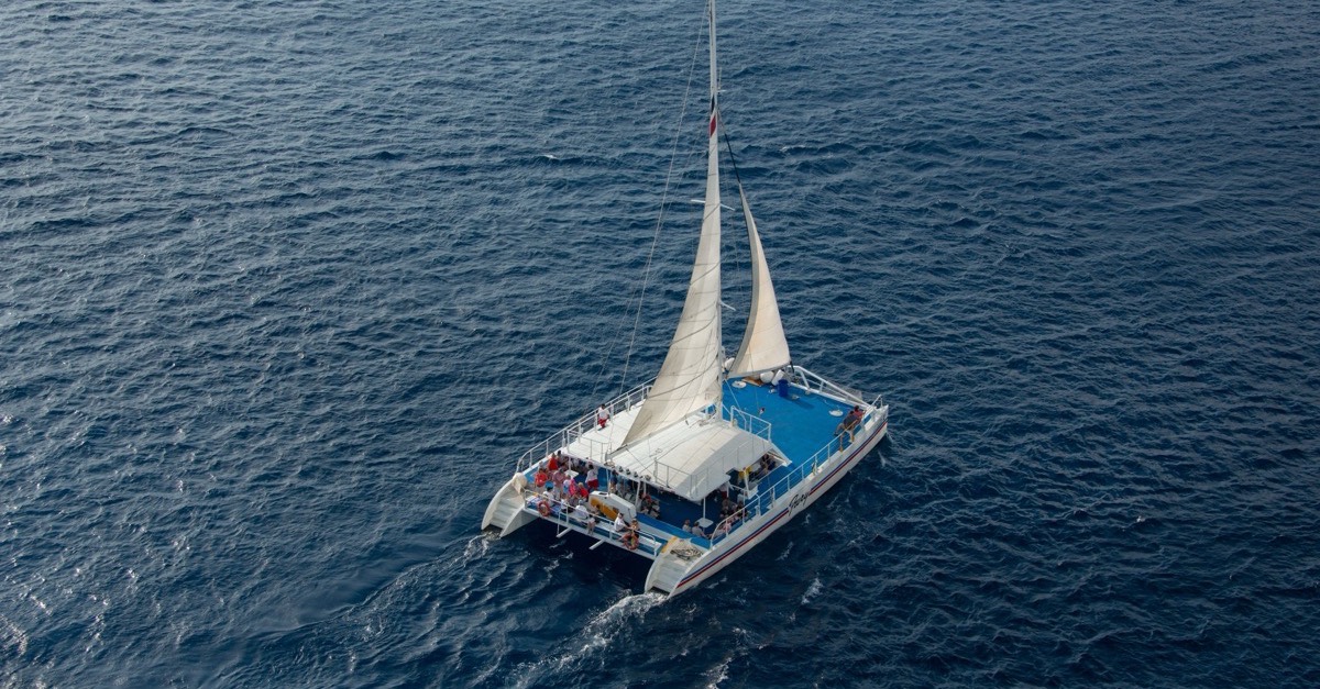 A sailing catamaran seen from above, gliding across calm blue water with passengers on deck